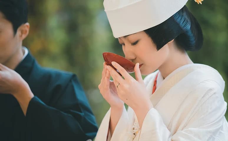 Bride in shiromuku and wataboshi drinking sacred sake from a red lacquer cup during the san-san-kudo ritual